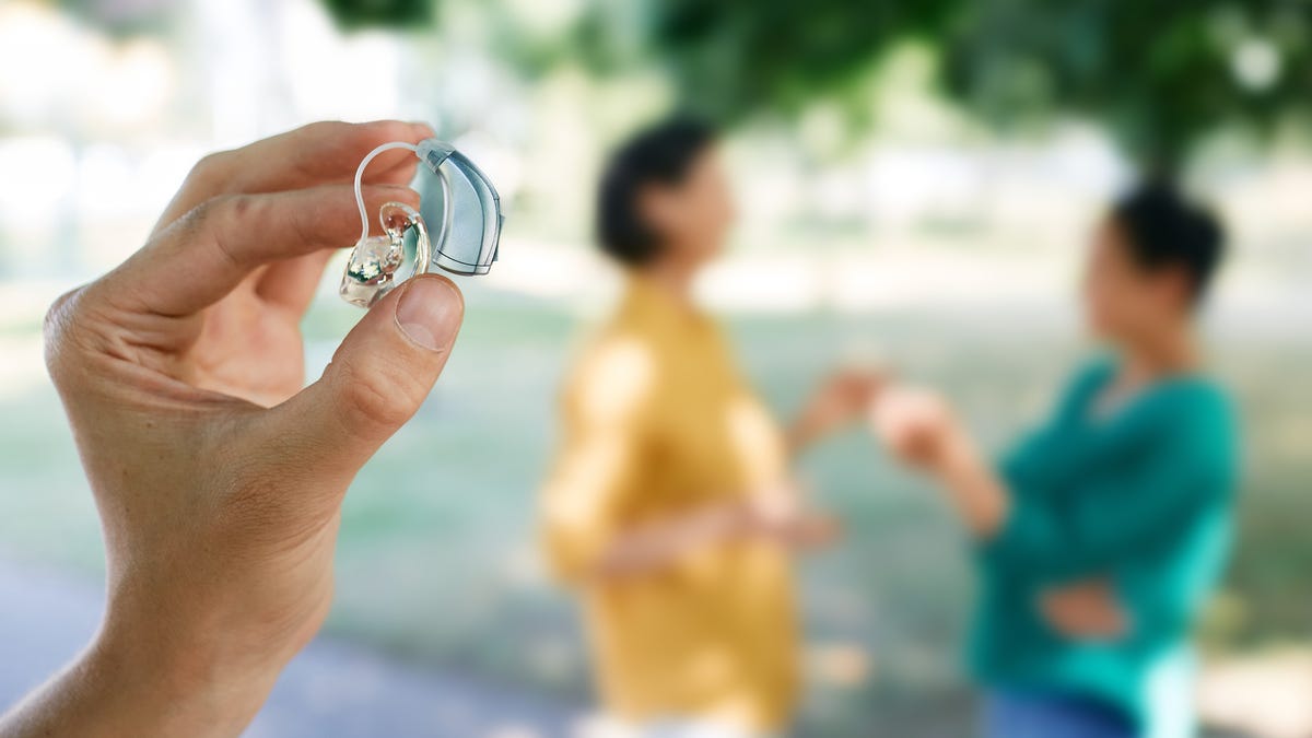 Person holding up a hearing aid as two people talk in the background