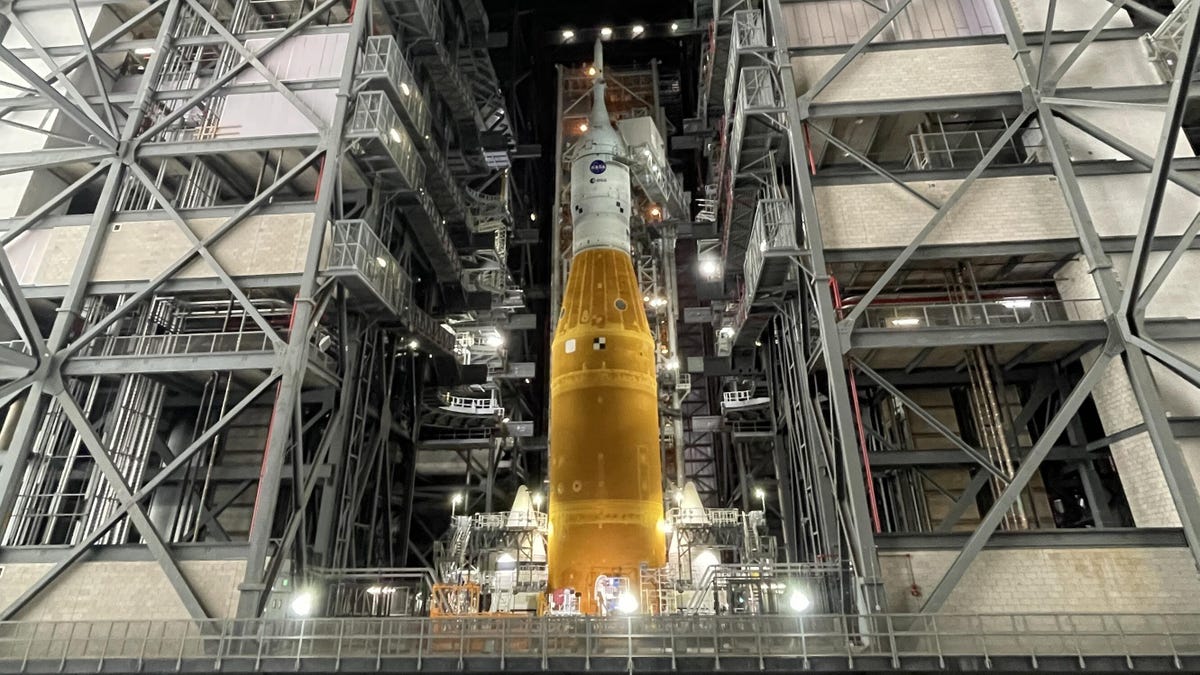 SLS and Orion, together a white capsule on top of an orange rocket surrounded by platforms inside a giant garage.