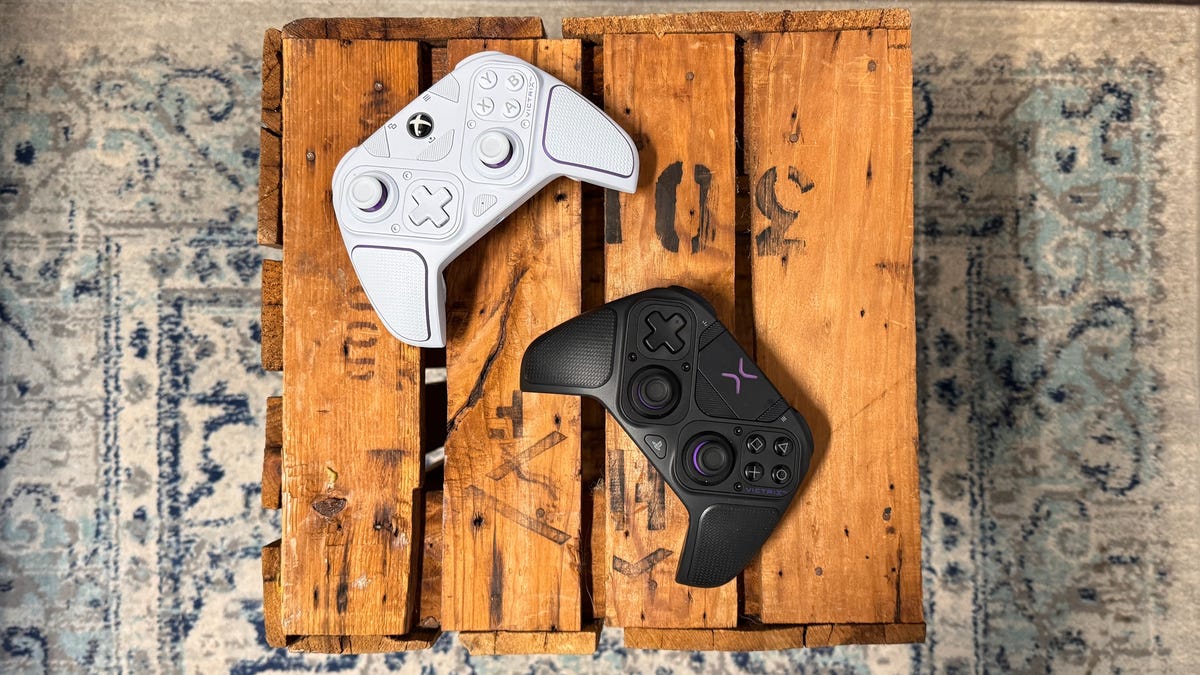 The PDP Victrix Pro BFG controllers for Xbox in white and for PS5 in black, sitting on a wood table.