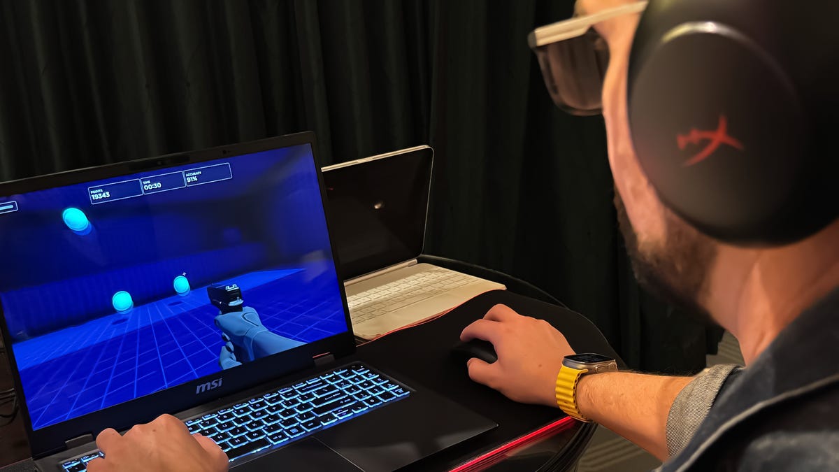 A man wears over-ear headphones while sitting at a desk with hands on a laptop and mouse.