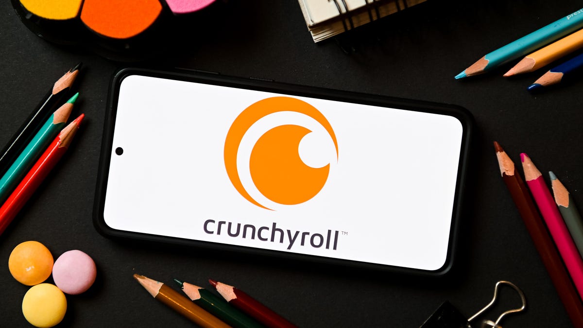 A phone showing the Crunchyroll logo. The phone is surrounded by pencils, notebooks and other office supplies