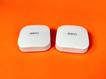 Two Eero mesh routers (small, white boxes that say "Eero" in black lettering on top) sit on an orange background.