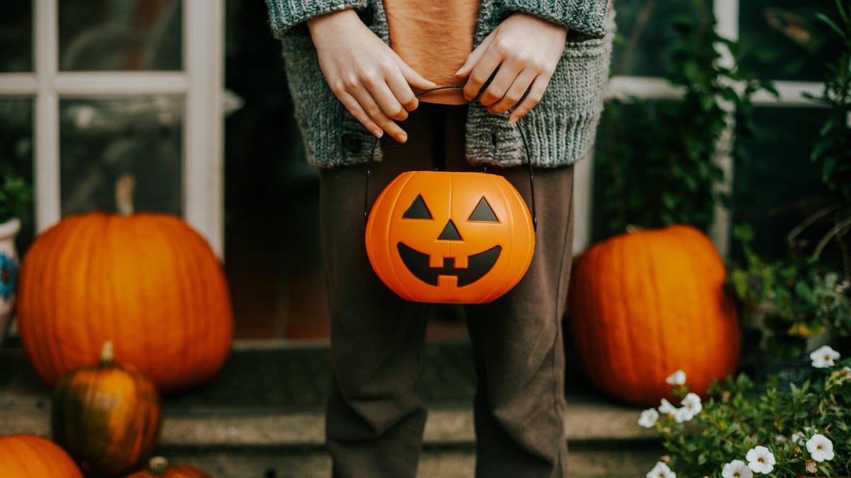 A person stands in front of pumpkins holding a jack o lantern treat bucket.