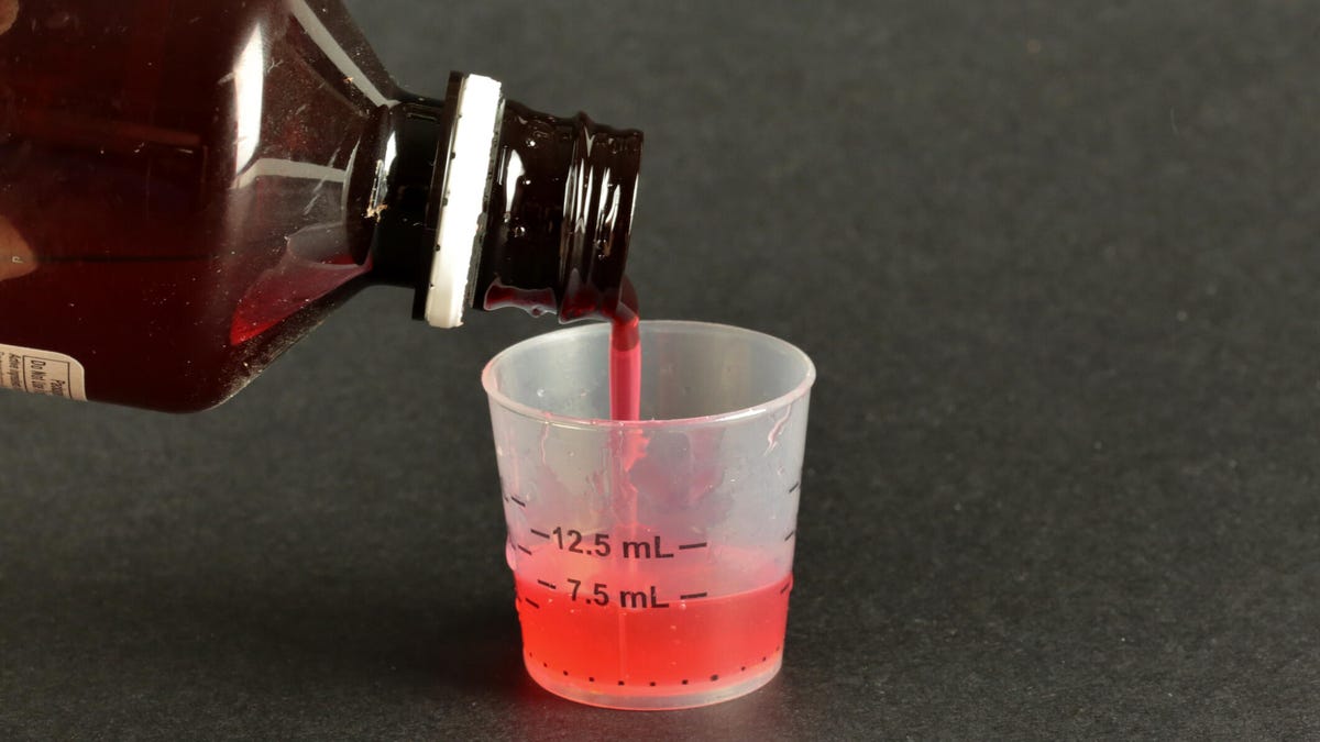 Red cough syrup being poured out of an amber bottle into a small measuring cut on a dark grey surface.
