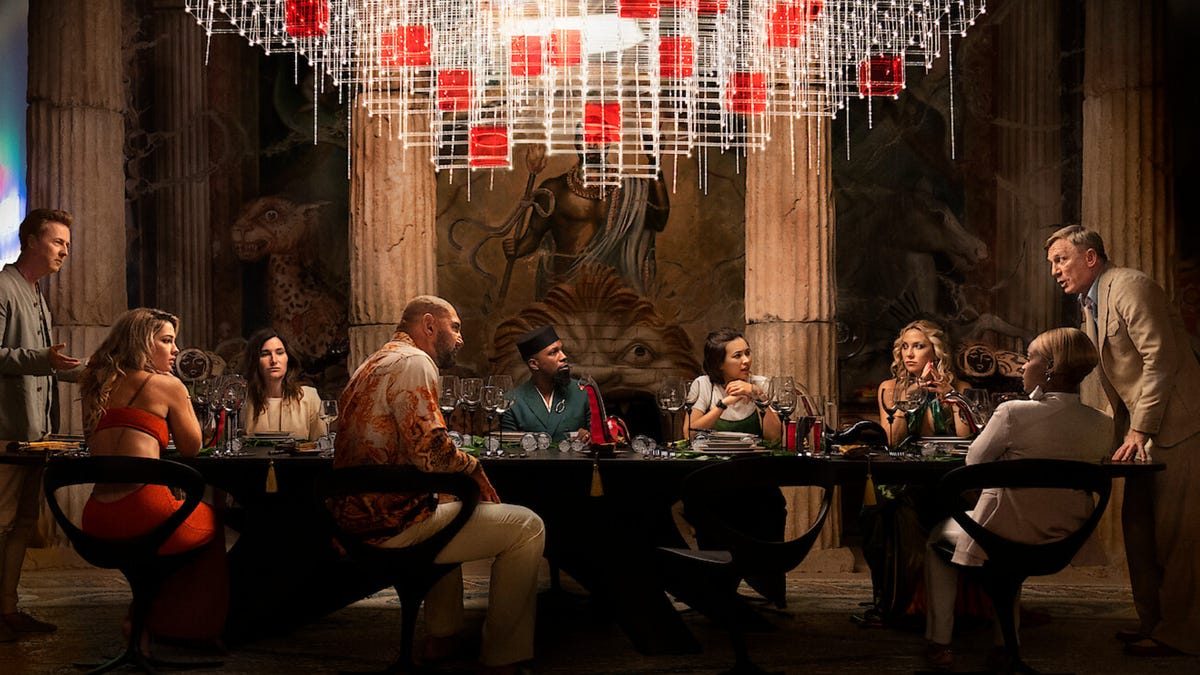 The characters in Glass Onion sit and stand at a long dinner table under an elaborate, geometric chandelier
