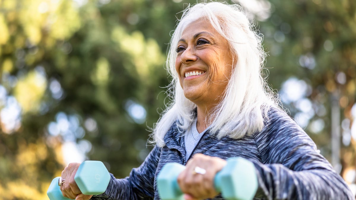 An older adult with shoulder-length white hair and a blue patterned sweatshirt lifting light-blue weights while surrounded by trees outside.