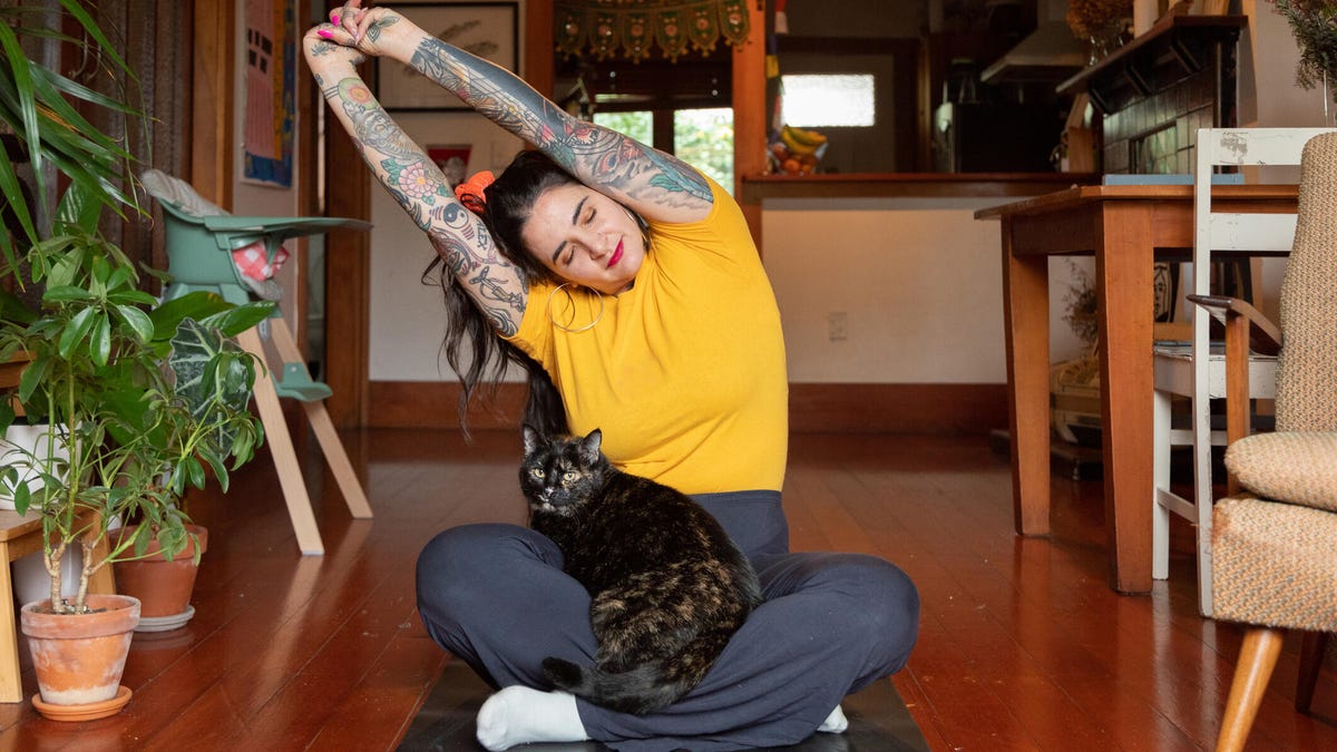A person with long brown hair, a yellow tee, dark blue pants and a cat on their lap stretching overhead while sitting on black mat on the floor in their home.