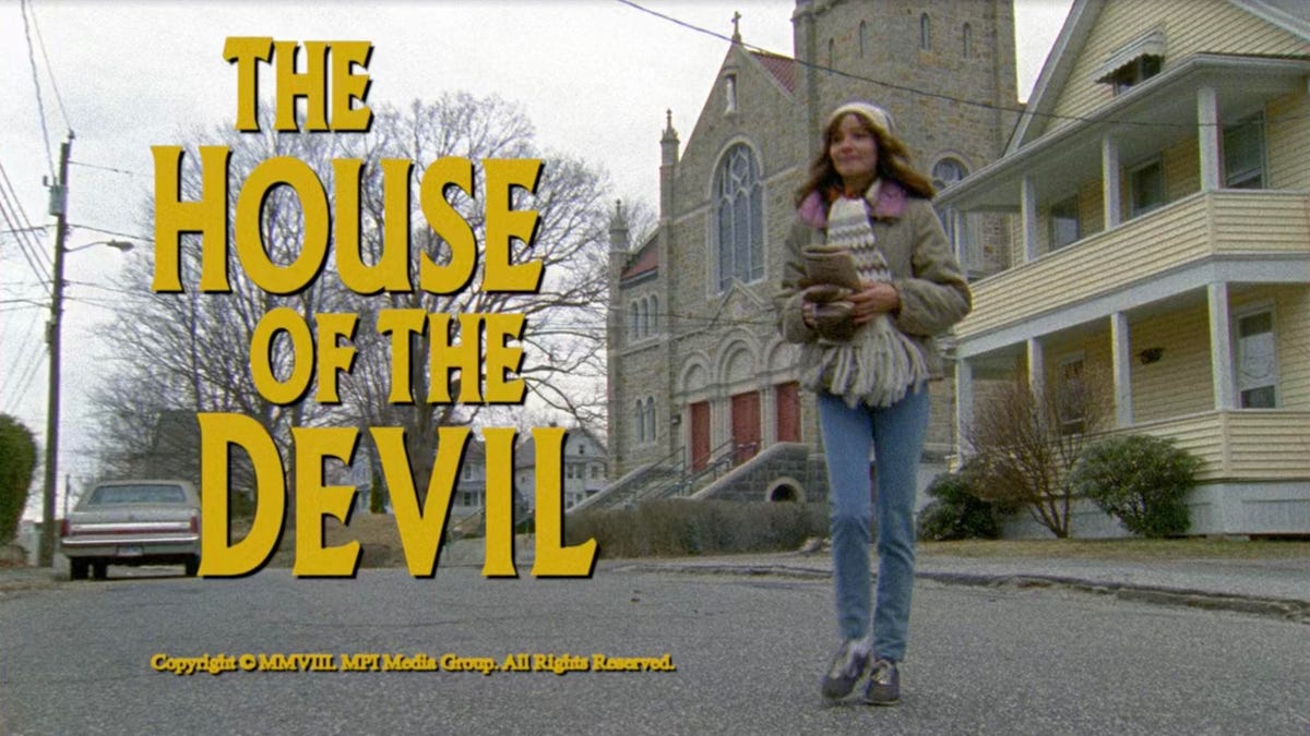 the title screen of the movie The House of the Devil, showing a young woman in winter clothing crossing a residential street with the title of the film superimposed on top