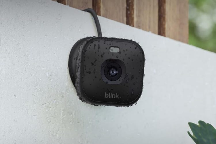 A black Blink mini camera on a white outside wall, wet with moisture.