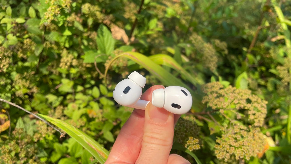 A hand holding a pair of AirPods Pro 2 against greenery