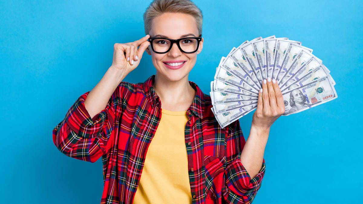 A person wearing a plaid shirt and glasses and holding a number of fanned out $100 bills.