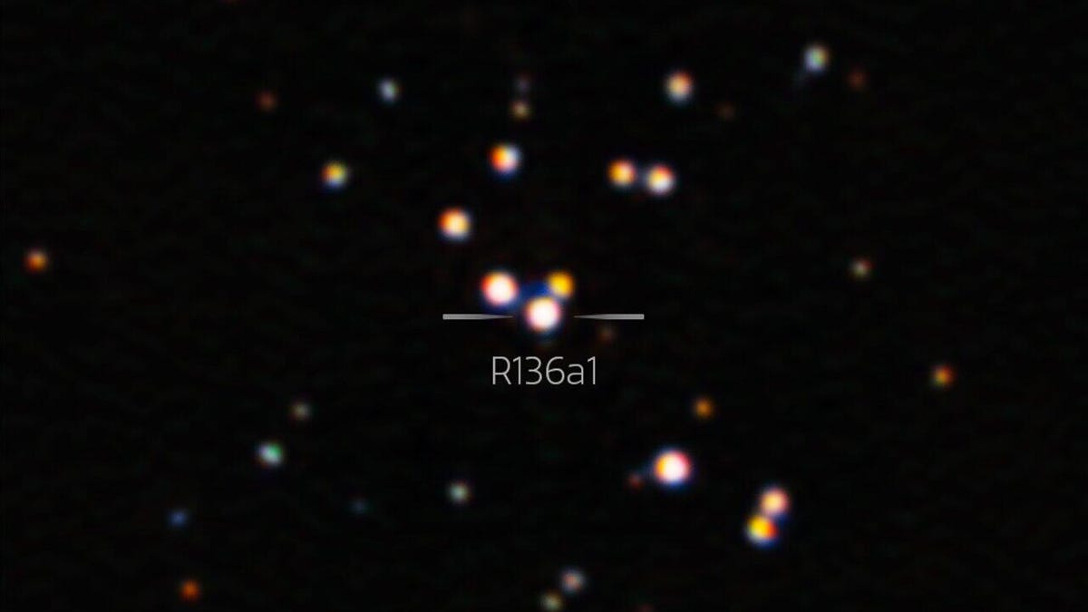 A dark background of space shows a few slightly blurry dots of light in various colors. They're concentrated toward the center, and the brightest one right in the middle is pinkish-red and represents R136a1.