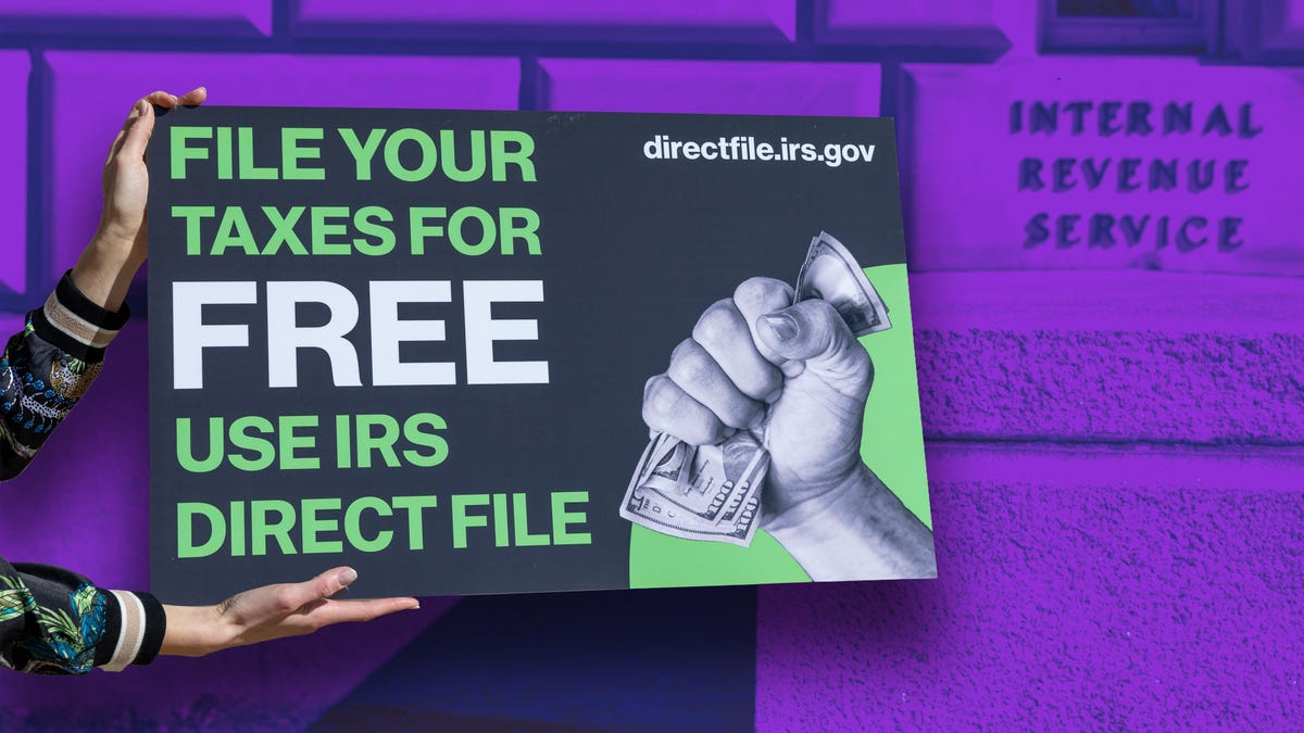 Hands holding a sign with "File your Taxes Free use IRS Direct File" against a purple colorized IRS building sign.