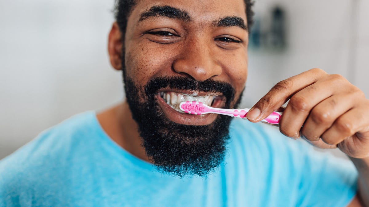 A person with black curly hair, a black beard and a bright blue tee brushing their teeth with a pink toothbrush.