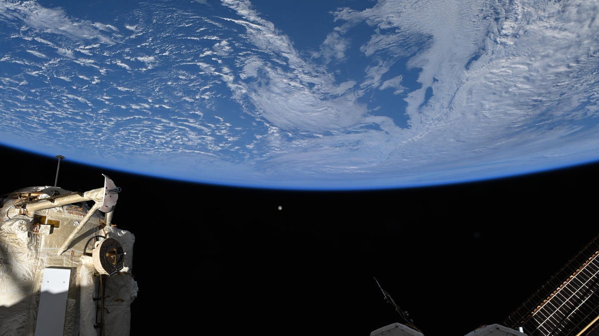 Earth seen from "below" in blue with clouds. Parts of the iSS in the lower frame. A full moon as a tiny dot in the center.