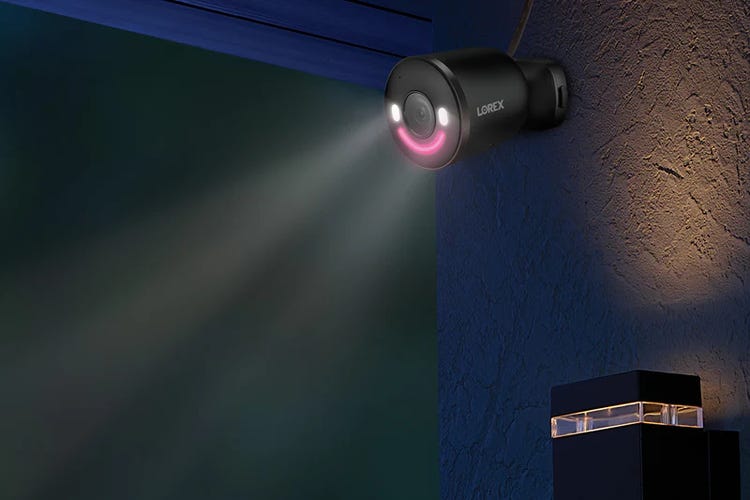 A Lorex 4K spotlight cam with lights on, posted on a stucco wall at night.