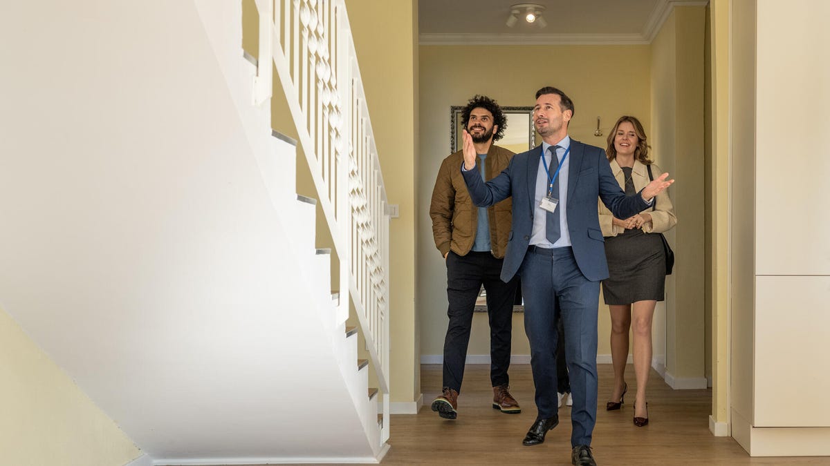 A real estate agent in a suit leads a couple through an empty house entryway with stairs.