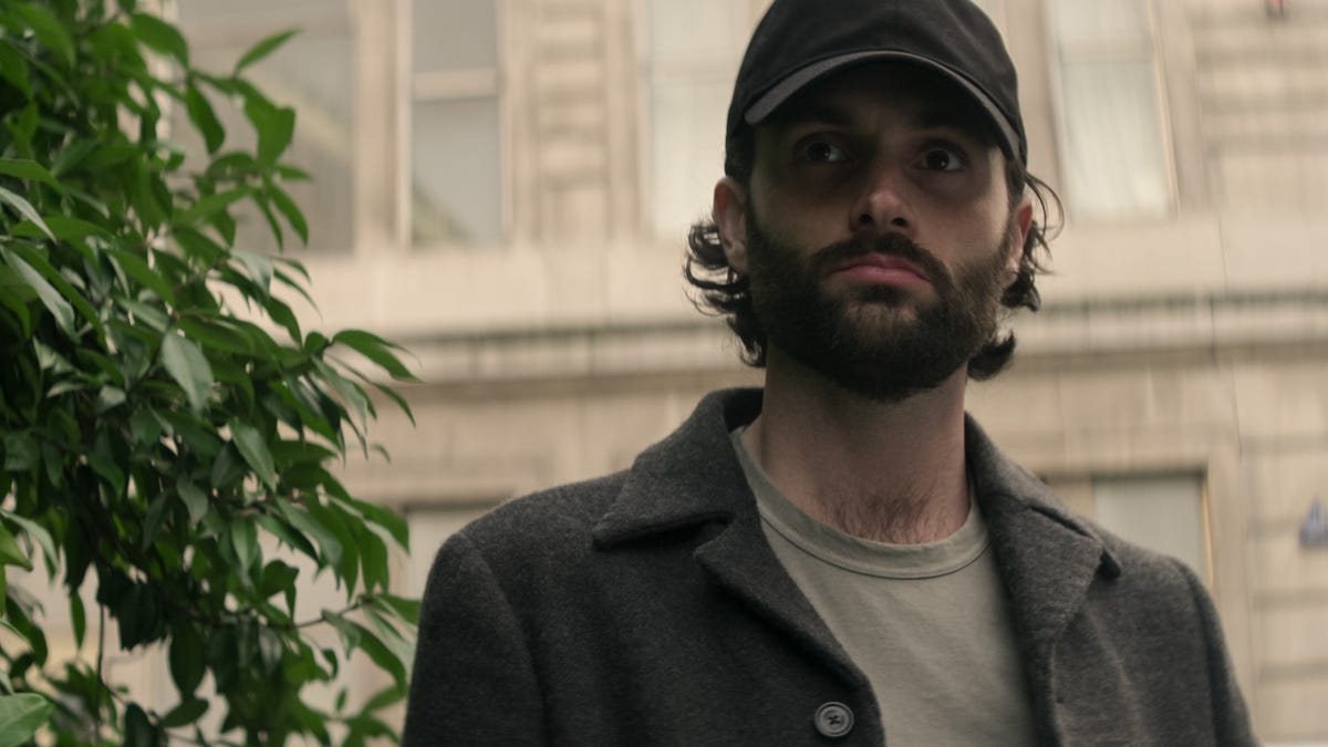 Midshot of Joe in a baseball cap standing in front of a building next to a tree, looking at something offscreen toward us