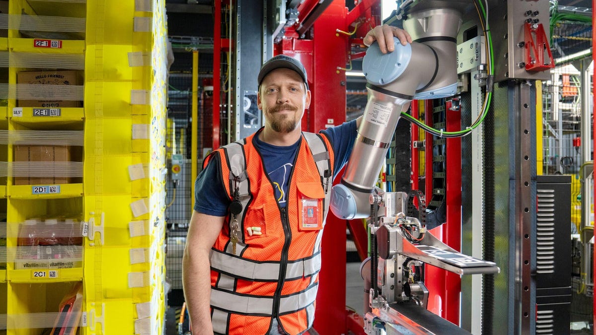 Human Amazon fulfillment center employee in orange vest stands next to Vulcan robot arm.
