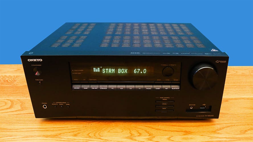 A black Onkyo TX NR6100 receiver on a wooden table with a blue background.