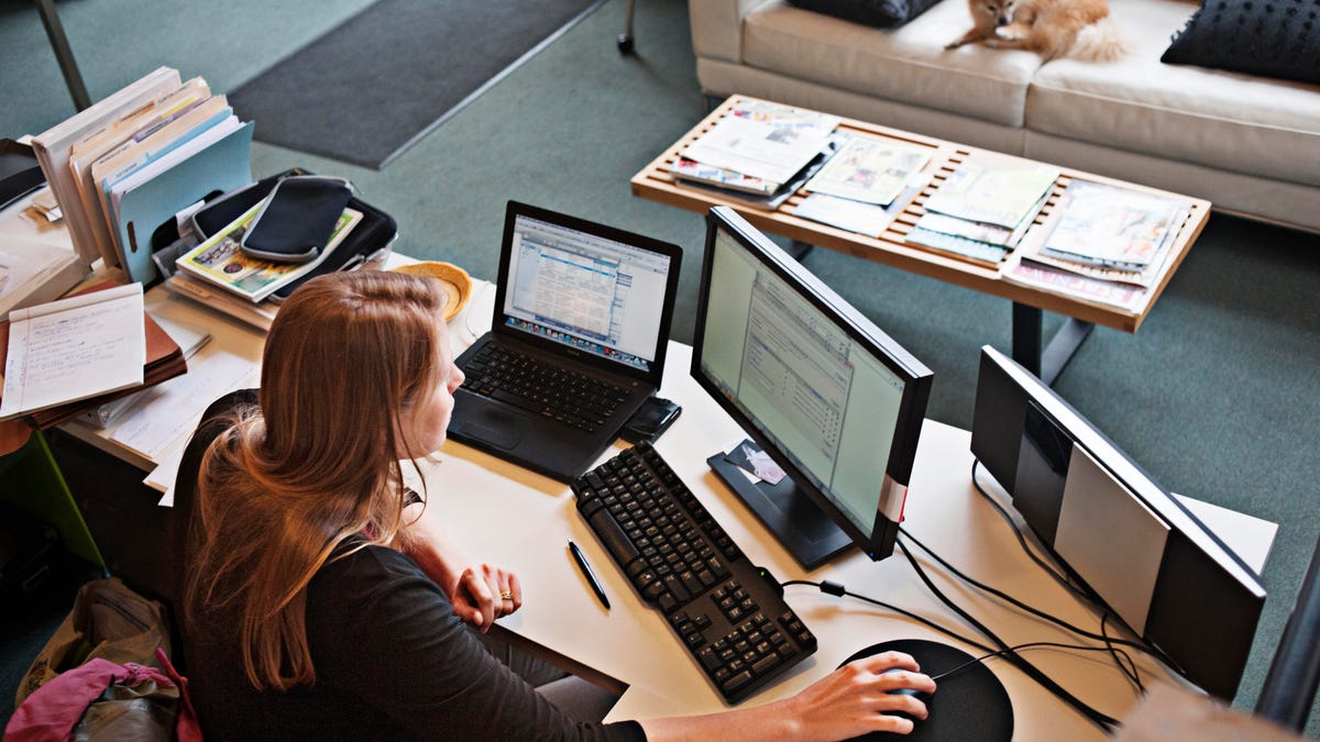 woman in home office with three computer screens and dog on couch