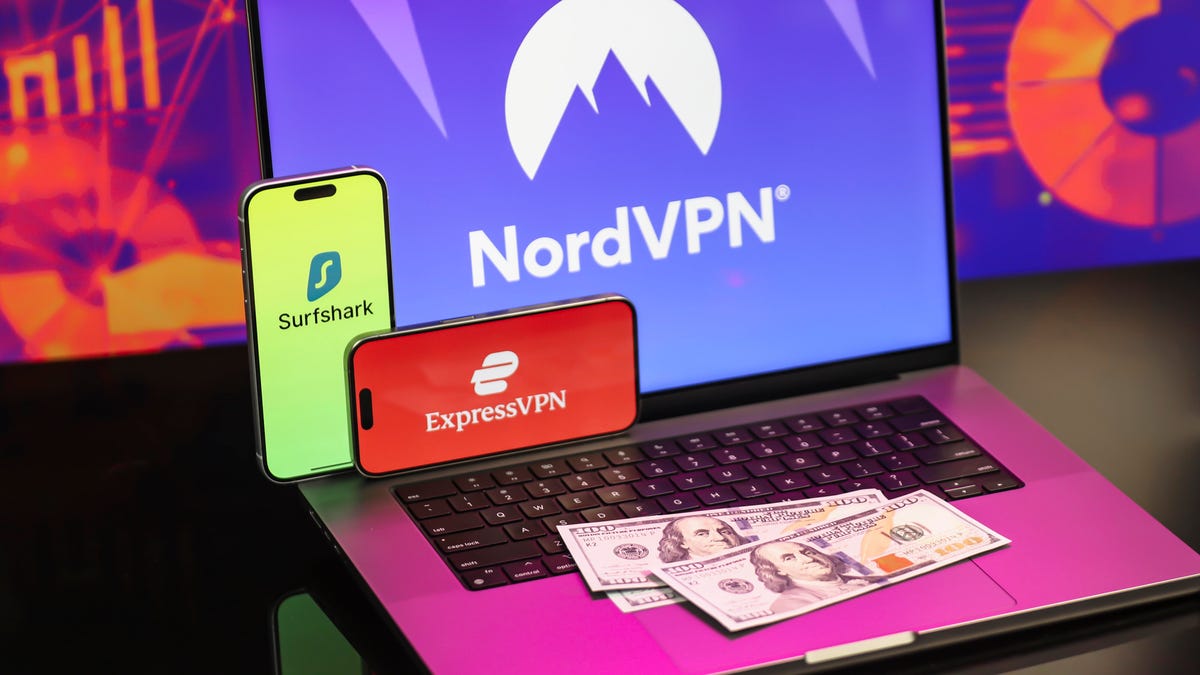 phone with a Surfshark logo displayed and another phone with ExpressVPN logo displayed, both perched on a laptop with NordVPN logo displayed; three one hundred dollar bills are also laid out on the laptop