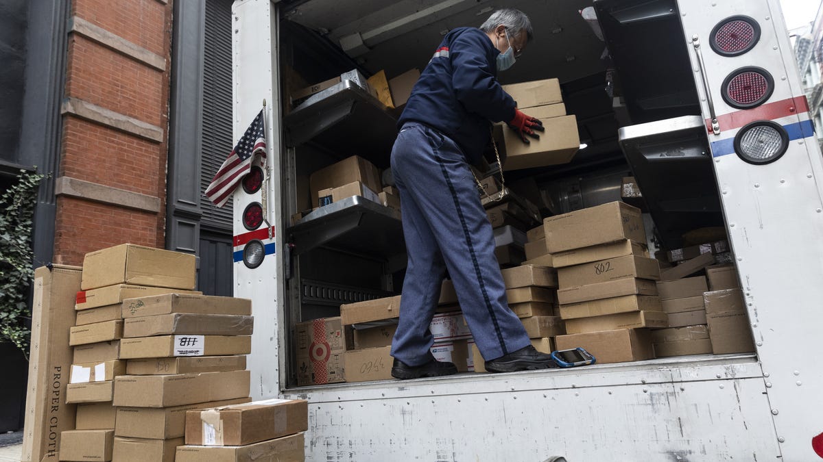 A postal worker stands in the back of an open USPS truck full of packages, with a pile of packages partially unloaded onto the ground below him.