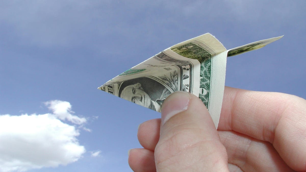 a hand holds a dollar bill folded into a paper airplane in front of a blue sky with white clouds in the lower left corner