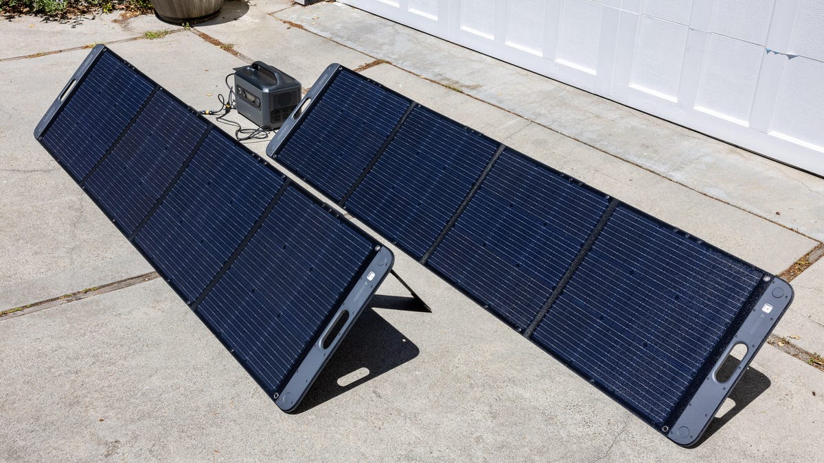 The two four-panel sections of Ugreen's PowerRoam 1200 Solar Generation products stand on a driveway, tilted toward the sun, with cables strung to the battery they're charging.