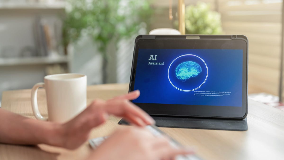 Hands type on a keyboard next to a mug and a tablet screen showing an AI assistant.