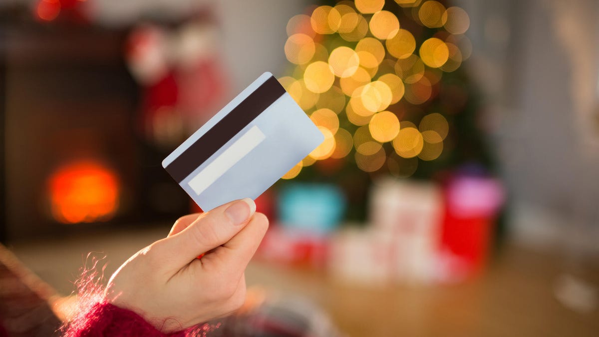 Someone holds a credit card in front of a blurred background of a Christmas tree with presents and a fireplace.
