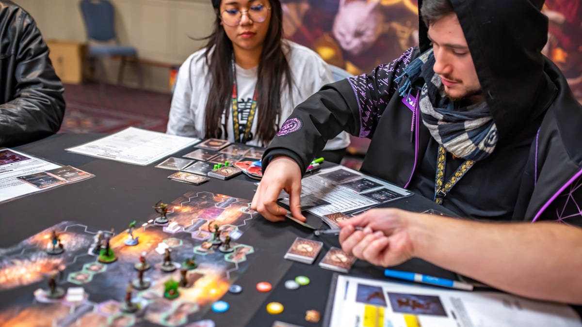 Several people gathered around a tabletop game move cards and miniatures to play.