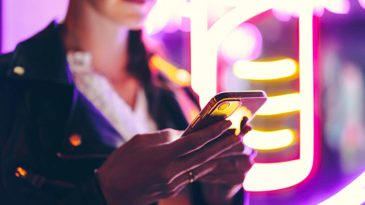 Woman using her smartphone at night, lit by purple neon lights gettyimages-1401071226