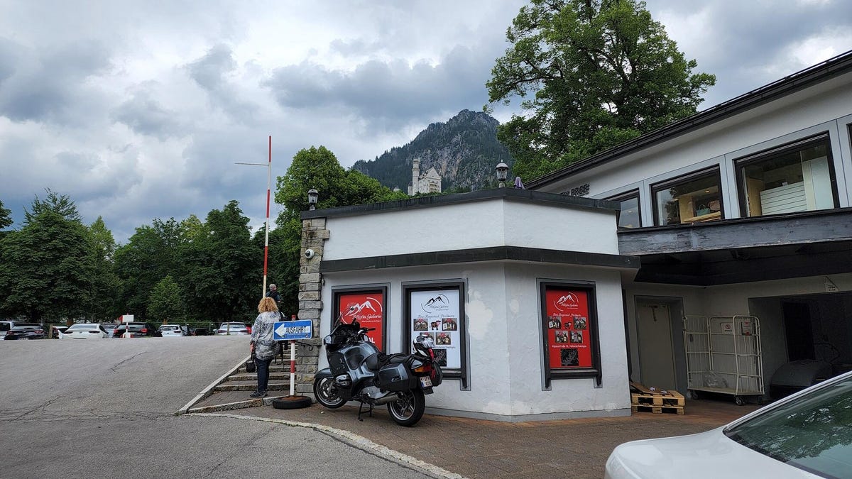 A view from the street with Neuschwanstein Castle in the distance. As the zoom increases, the castle comes into view: baroque in white stone that shines brightly against the lush green forest around it.