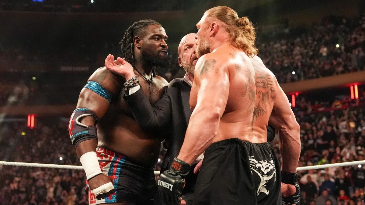 A Black wrestler and white wrestler face off in a ring with a man in a suit stepping between them.