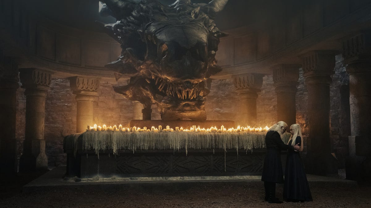 Wide shot of King Viserys talking to Princess Rhaenyra in a crypt overlooked by a giant stone dragon's head.