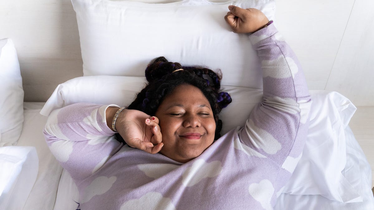 A person with pulled-back dark brown hair wearing a light purple shirt with white clocks waking up happy in bed.