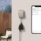 An Amazon Basics outdoor smart plug on a gray wall next to a patio with the app shown beside it.