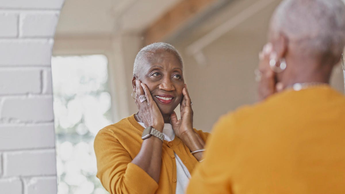 A person in a yellow shirt touches her skin while looking in the mirror