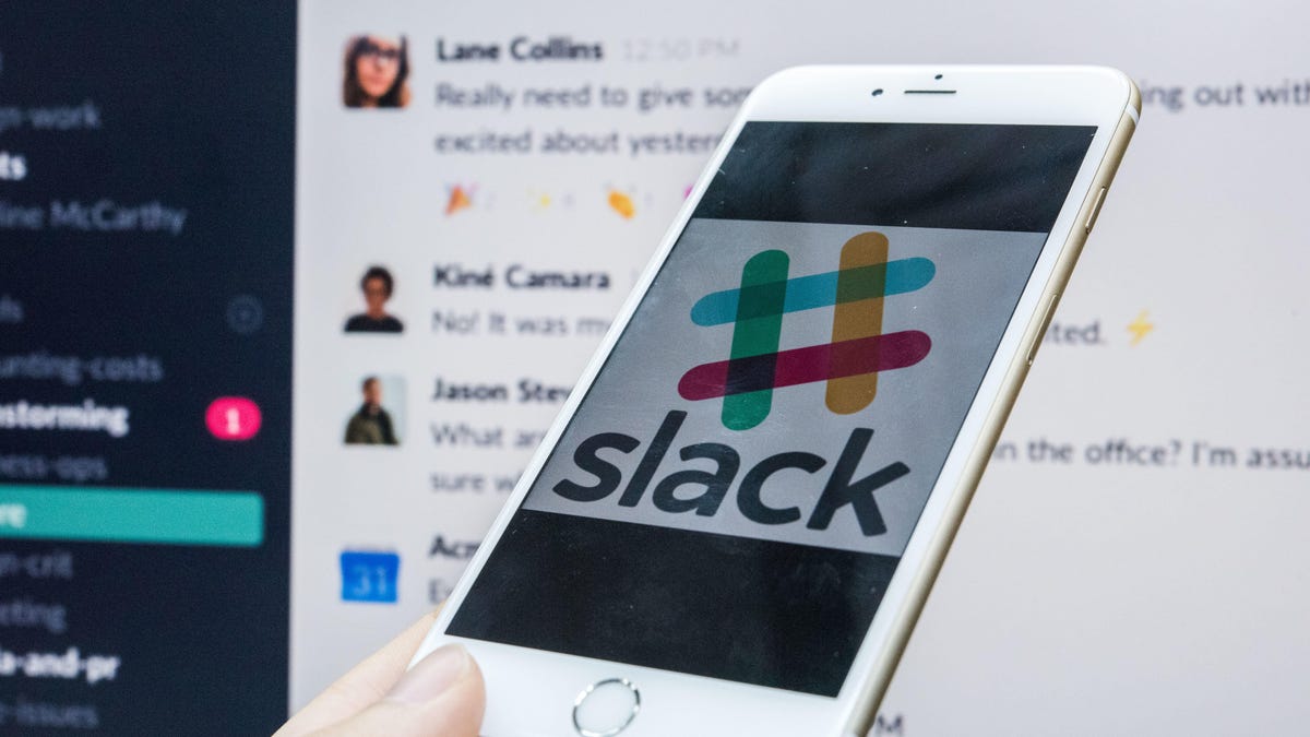 An iPhone shows the Slack logo on its screen, in front of a desktop screen showing a Slack channel.