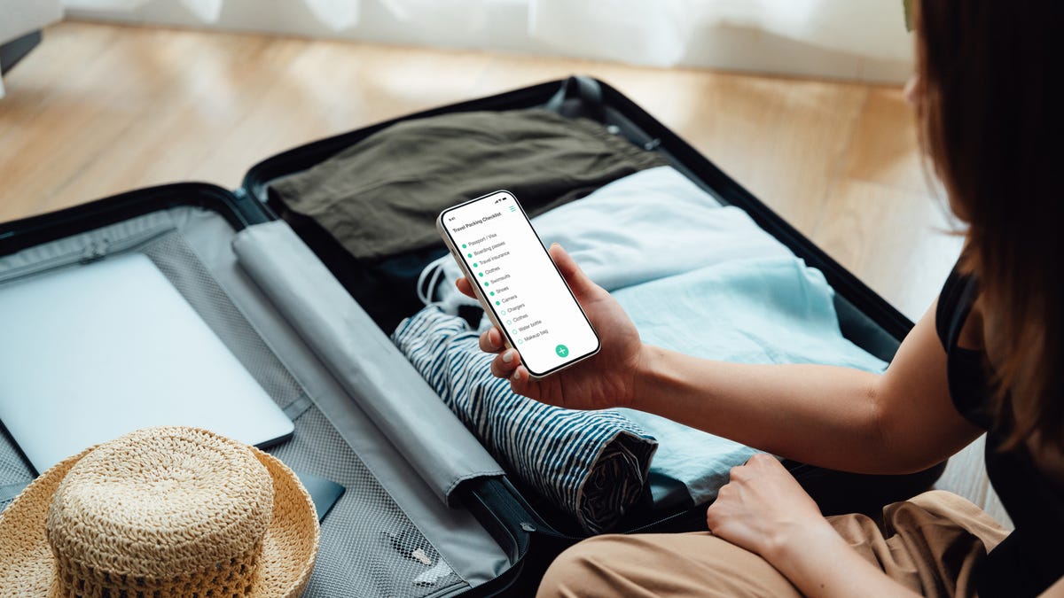 an over the shoulder view of a woman looking at a checklist on her phone while packing a suitcase