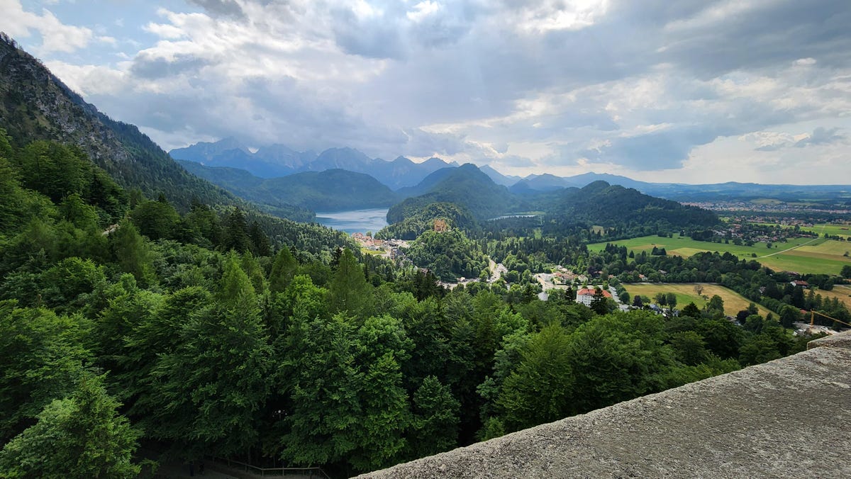 A view of the lush tree-filled valley from the balcony of Neuschwanstein Castle, with the zoom showing closer views of the opposite Hohenschangau castle at the mountain foothills with cloud-wracked open sky above.
