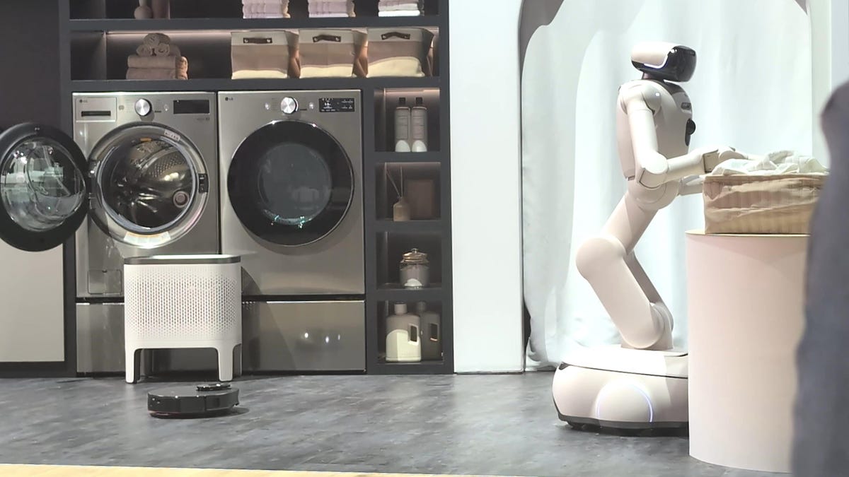A humanoid robot folds laundry, with a washer and dryer in the background