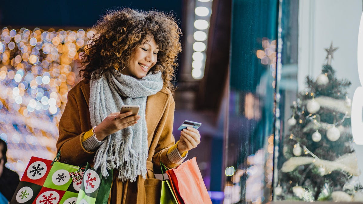 A woman holds numerous shopping bags while looking at her credit card outside of another store.