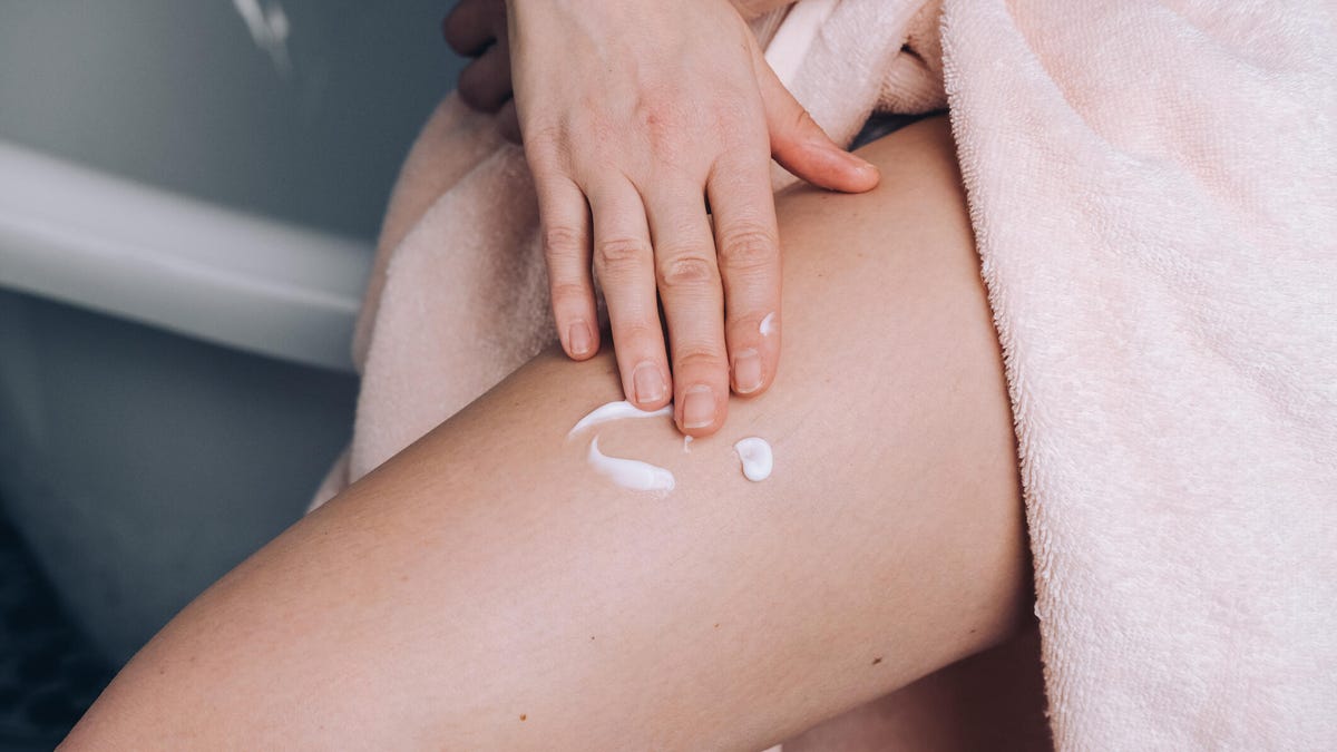 A person wearing a light pink towel using their hand to rub in white cream on their leg.