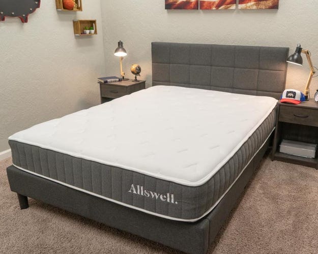 An Allswell hybrid mattress rests on a bedframe between two side tables with the brand label clearly shown on the side of the mattress.