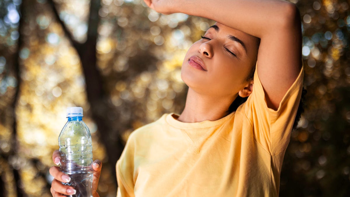 A person is a yellow tee sweating and drinking water outside with their arm over their head.