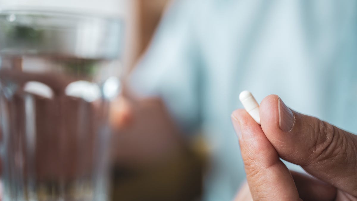 A hand holding a small white capsule with a glass of water in the background.