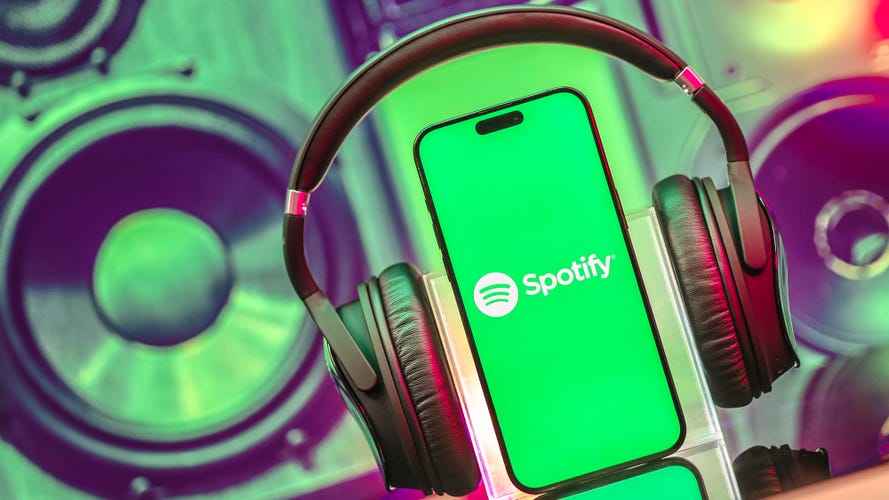 Headphones are placed on an upright cellphone, as if the cell is listening through the headphones. The Spotify music logo is on the cell's screen.