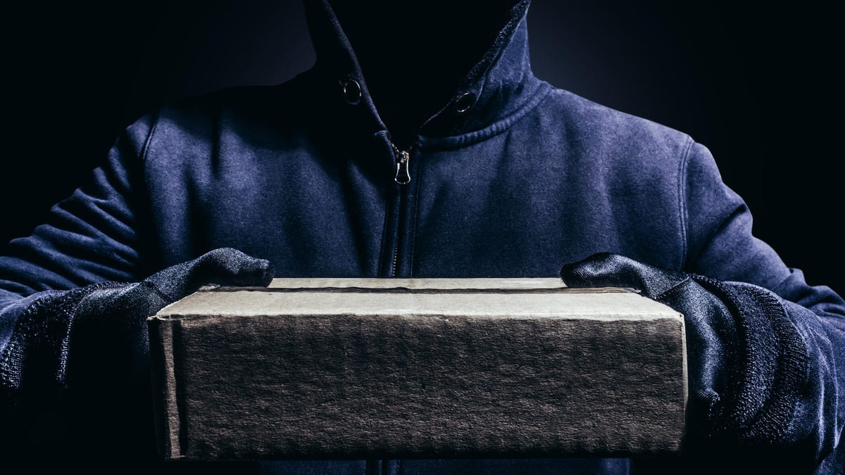 A hooded figure in gloves lifts a brown package.